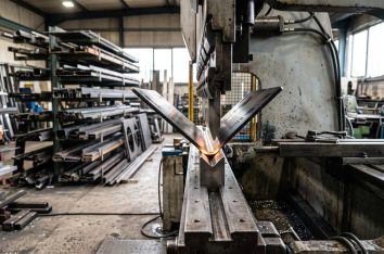 Press brake forming a laser-cut steel blank, the part mid-bend between punch and die, fabrication shop with racked...