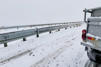 Salted snowy road shoulder with slush spray on a steel guardrail and a truck bumper in winter
