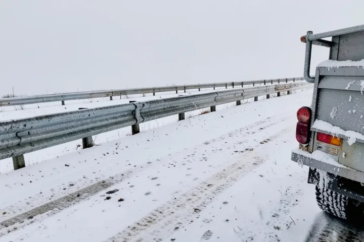Salted snowy road shoulder with slush spray on a steel guardrail and a truck bumper in winter